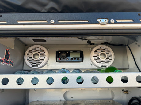 Custom RV rental setup features a Fusion stereo, speakers, and water bottles in a lit storage area. Good vibes!