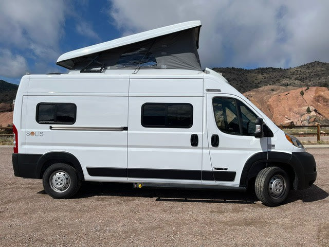 Side view of a white Winnebago Solis pop-top rv rental van parked on gravel in a mountainous area.
