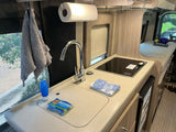 An RV rental's functional kitchen featuring a sink, cooktop, paper towels, and cleaning supplies. Ideal for on-the-go meals.