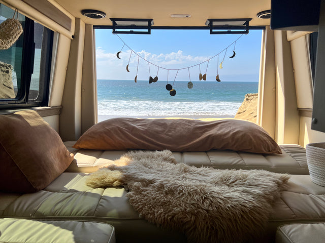 Relaxing RV rental interior with a cozy bed, pillows, and sheepskin rug overlooking a stunning ocean view and beach.