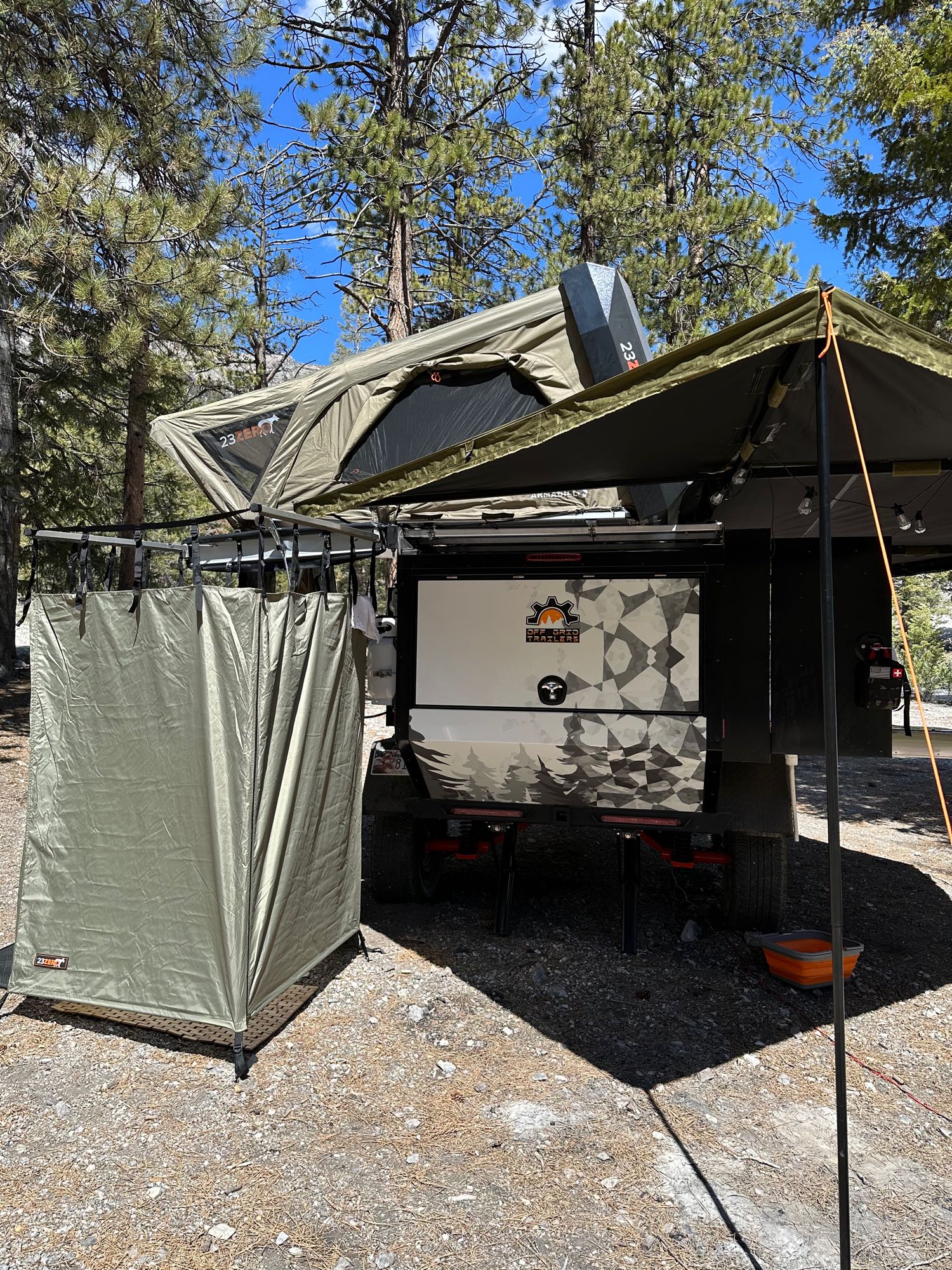 An olive green off-road camping trailer is set up with a rooftop tent, side awning, and privacy tent, perfect for an RV rental.