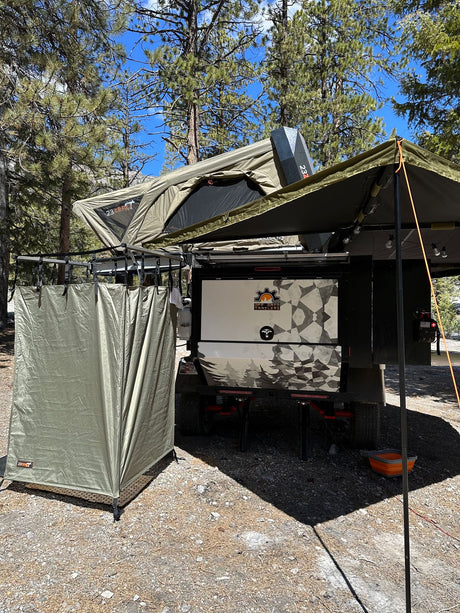 An olive green off-road camping trailer is set up with a rooftop tent, side awning, and privacy tent, perfect for an RV rental.