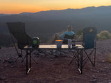 Scenic RV rental spot at dusk: person relaxing in a chair, viewing mountains. Table with camp gear in foreground.