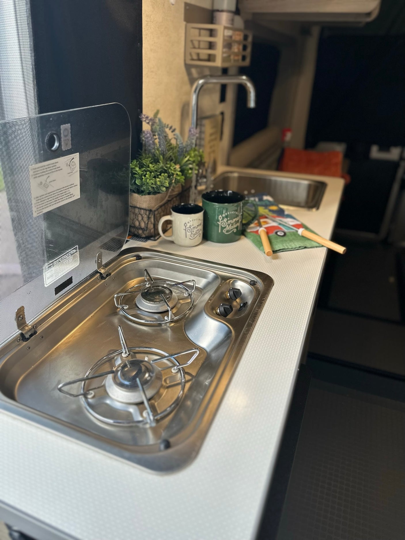 Compact RV rental kitchen featuring a two-burner stove, sink, and countertop with accessories. Ideal for cooking on the road.