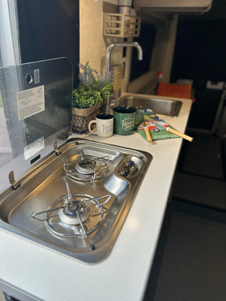 Compact RV rental kitchen featuring a two-burner stove, sink, and countertop with accessories. Ideal for cooking on the road.