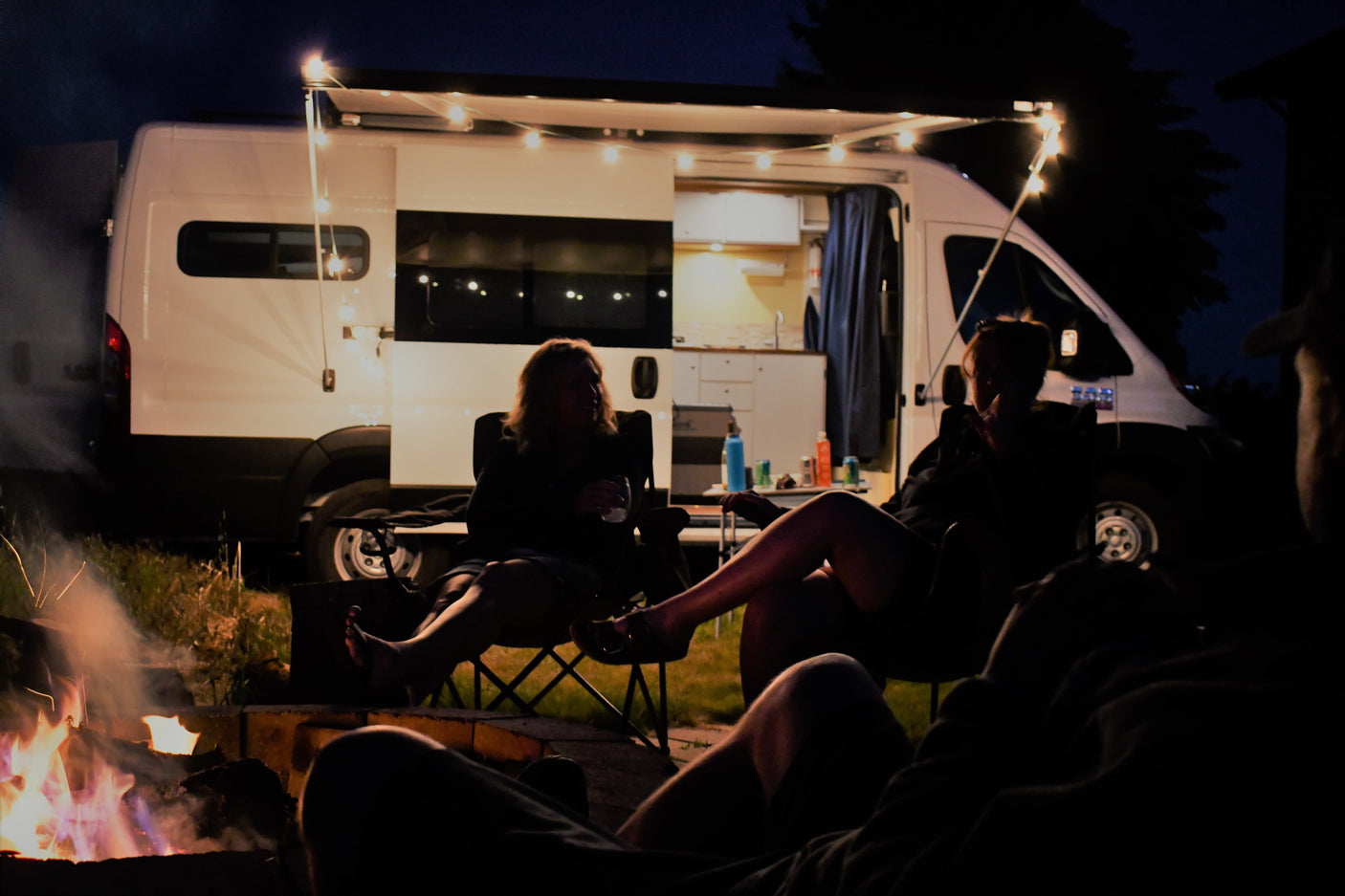 Nighttime RV rental scene: Friends relax by a warm campfire next to a white camper van with string lights.