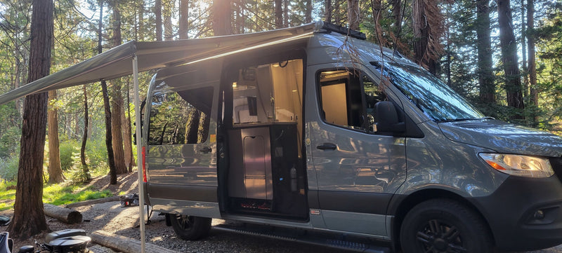 This RV rental camper van, with an extended awning and visible interior kitchen, is parked in a sunny forest campsite. — photo 6