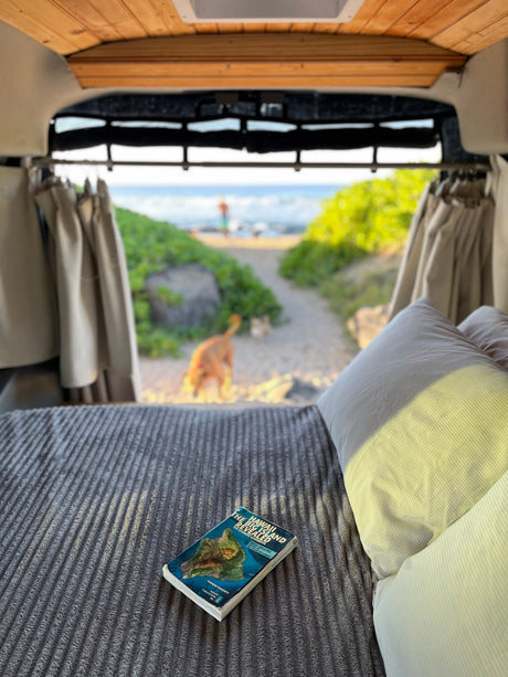 Cozy RV rental bed with a Hawaii guidebook. Looking out to a beach path, a dog, and a person by the ocean.