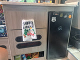 RV rental interior: organized cabinets, a fridge with travel stickers, & a "Happy Camper" towel. Cozy camper amenities.