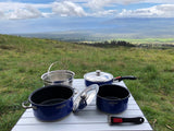 RV rental outdoor cooking: blue pots, pans, strainer on a table with a stunning ocean view from a grassy hill. Camp kitchen ready.
