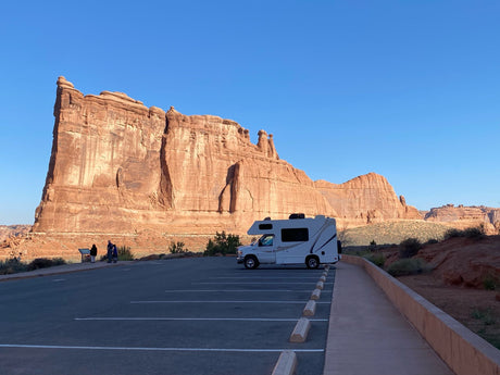 An RV rental sits in a sunlit parking lot, showcasing majestic red rock formations in a stunning desert national park.