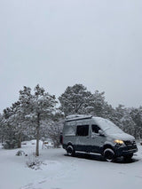 Snow-covered gray Sprinter RV rental van nestled in a serene winter forest. A peaceful travel scene.