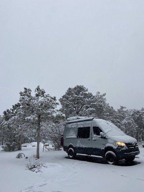 Snow-covered gray Sprinter RV rental van nestled in a serene winter forest. A peaceful travel scene.
