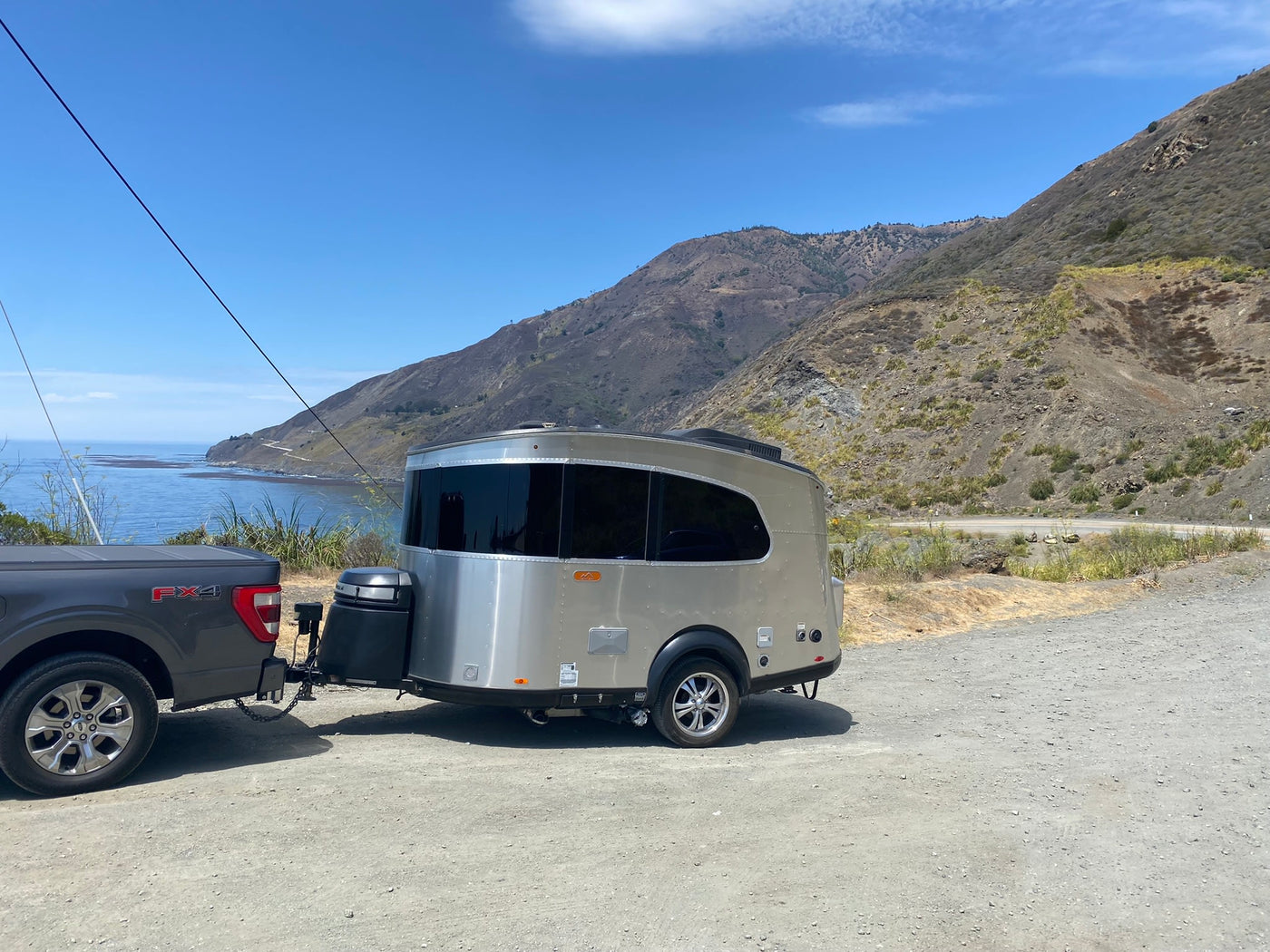A sleek, silver travel trailer, like an RV rental, is hitched to a truck on a gravel pull-off overlooking a scenic coast & mountains. — photo 11