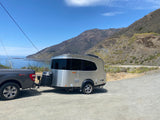 A sleek, silver travel trailer, like an RV rental, is hitched to a truck on a gravel pull-off overlooking a scenic coast & mountains.