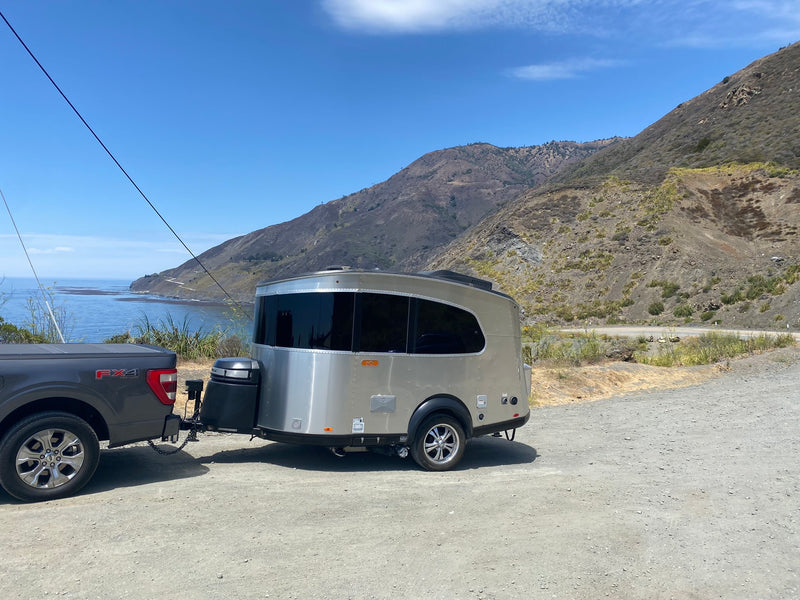 A sleek, silver travel trailer, like an RV rental, is hitched to a truck on a gravel pull-off overlooking a scenic coast & mountains. — photo 11