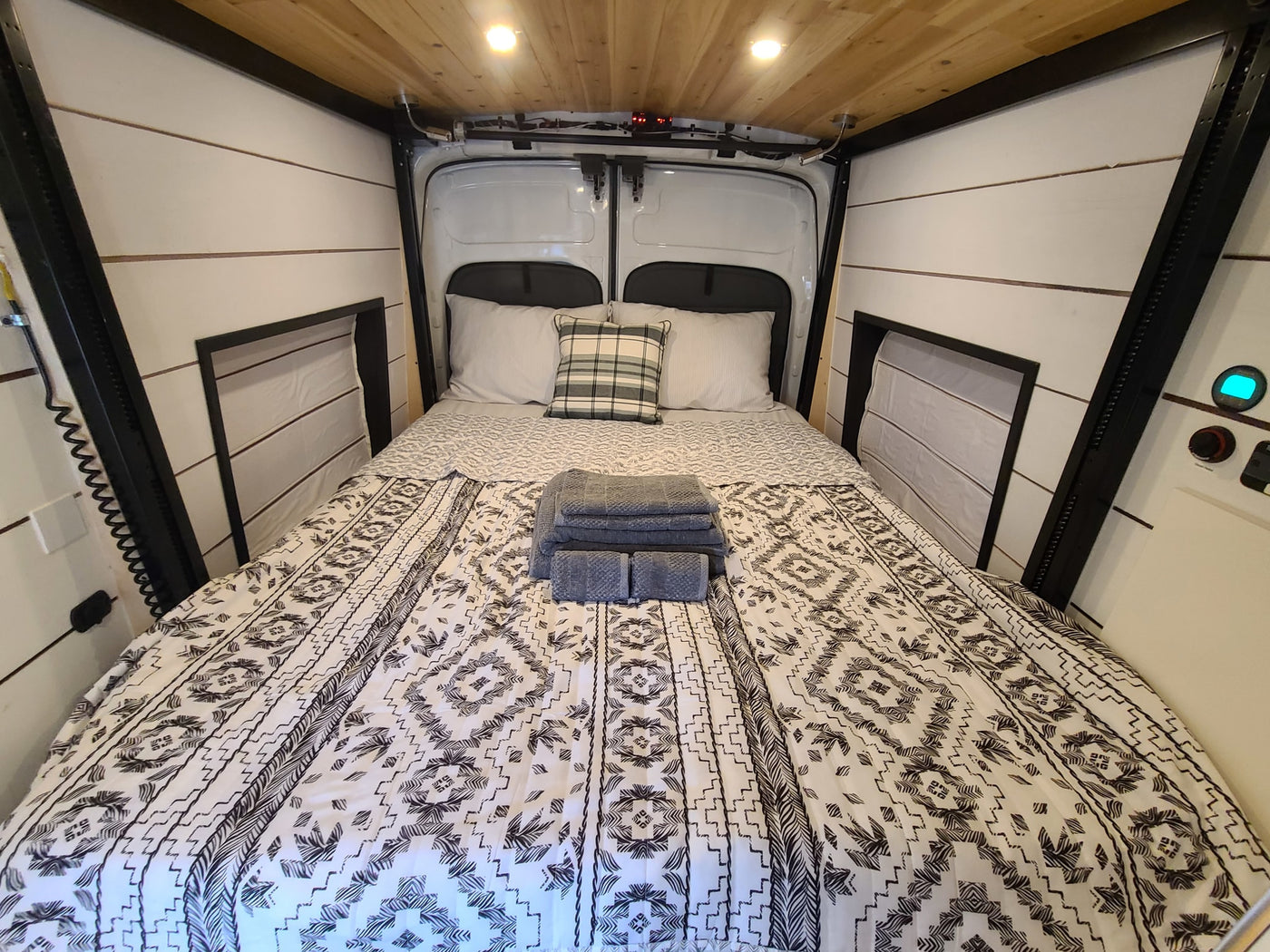 Cozy RV rental sleeping area: Bed with patterned black/white bedding, pillows, fresh towels, wood ceiling, & warm lighting. — photo 7