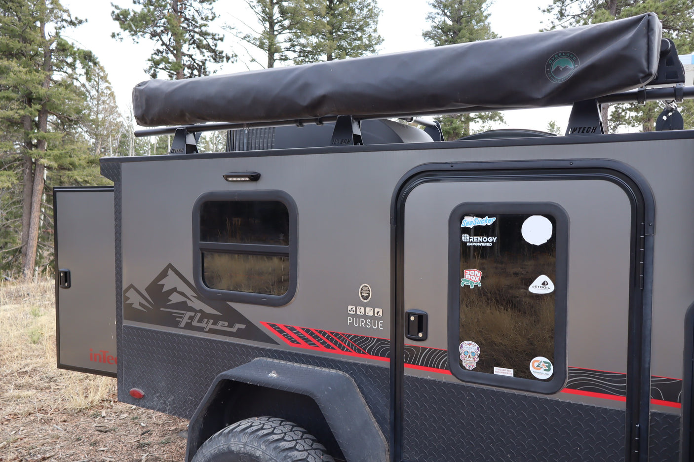 A gray InTech Flyer Pursuit **rv rental** **off-road camper** with an awning, adventure stickers, and rugged tires, parked in a wooded area.
