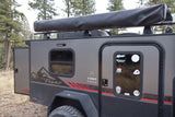 A gray InTech Flyer Pursuit **rv rental** **off-road camper** with an awning, adventure stickers, and rugged tires, parked in a wooded area.
