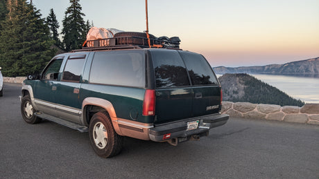 A green Chevrolet Suburban, outfitted for an outdoor adventure or rv rental, overlooks a tranquil lake and mountains at sunset.
