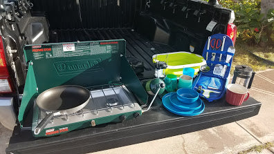 Setup for an RV rental adventure: Coleman stove, pan, dishes, and kitchen tools displayed on a truck tailgate.