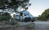 Modern RV rental camper van with open door parked in a natural, tree-lined setting, showing its light-wood interior.