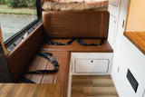 Spacious RV rental interior: L-shaped brown leather seating, window to wet road, white cabinetry, wood floor.
