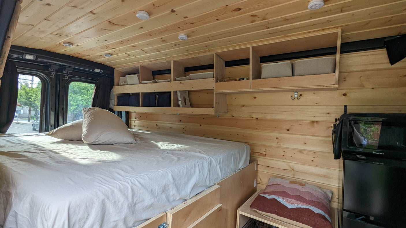 Cozy RV rental interior: wood-paneled walls/ceiling, comfortable bed with white linens, overhead storage, and compact kitchen.