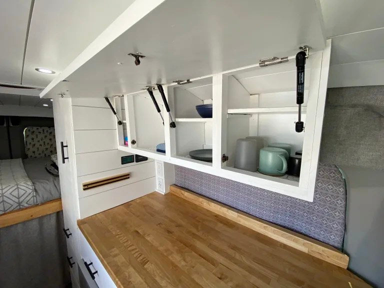 This RV rental's modern van conversion features open white overhead cabinets, a light wood counter, and cozy bed. — photo 15