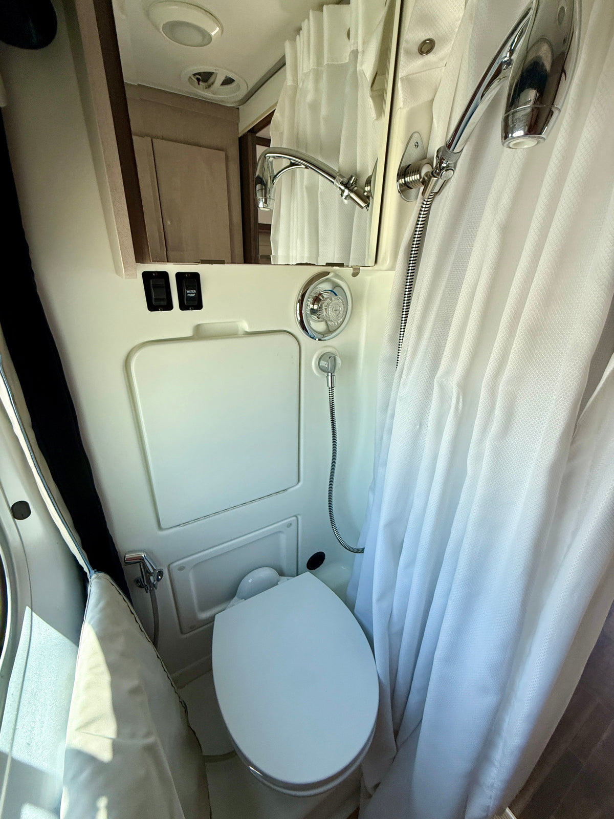 Compact RV rental bathroom featuring a white toilet, shower with curtain, mirror reflecting a faucet, and water pump switch. — photo 7