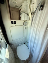 Compact RV rental bathroom featuring a white toilet, shower with curtain, mirror reflecting a faucet, and water pump switch.