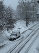 An RV rental is snowed in by a winter storm, parked roadside with a foot of snow, as tire tracks mark the street.