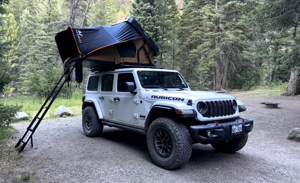  ready for off-road camping. An adventurous rv rental option in the forest.|White Jeep Wrangler with a rooftop tent set up in a forest. Experience outdoor camping with this unique RV rental.|White Jeep Wrangler Rubicon 4x4 with a rooftop tent. An ideal RV rental for rugged camping and outdoor adventure.|Experience adventure with this white Jeep Rubicon featuring a rooftop tent — photo 2