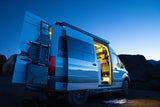 Night camping with a Sandy Vans camper van rv rental. Open doors reveal a cozy, illuminated interior and rear gear rack under a twilight sky.