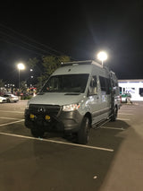 This custom light grey Mercedes-Benz Sprinter adventure van, ideal for RV rental, is parked in a bright nighttime lot.