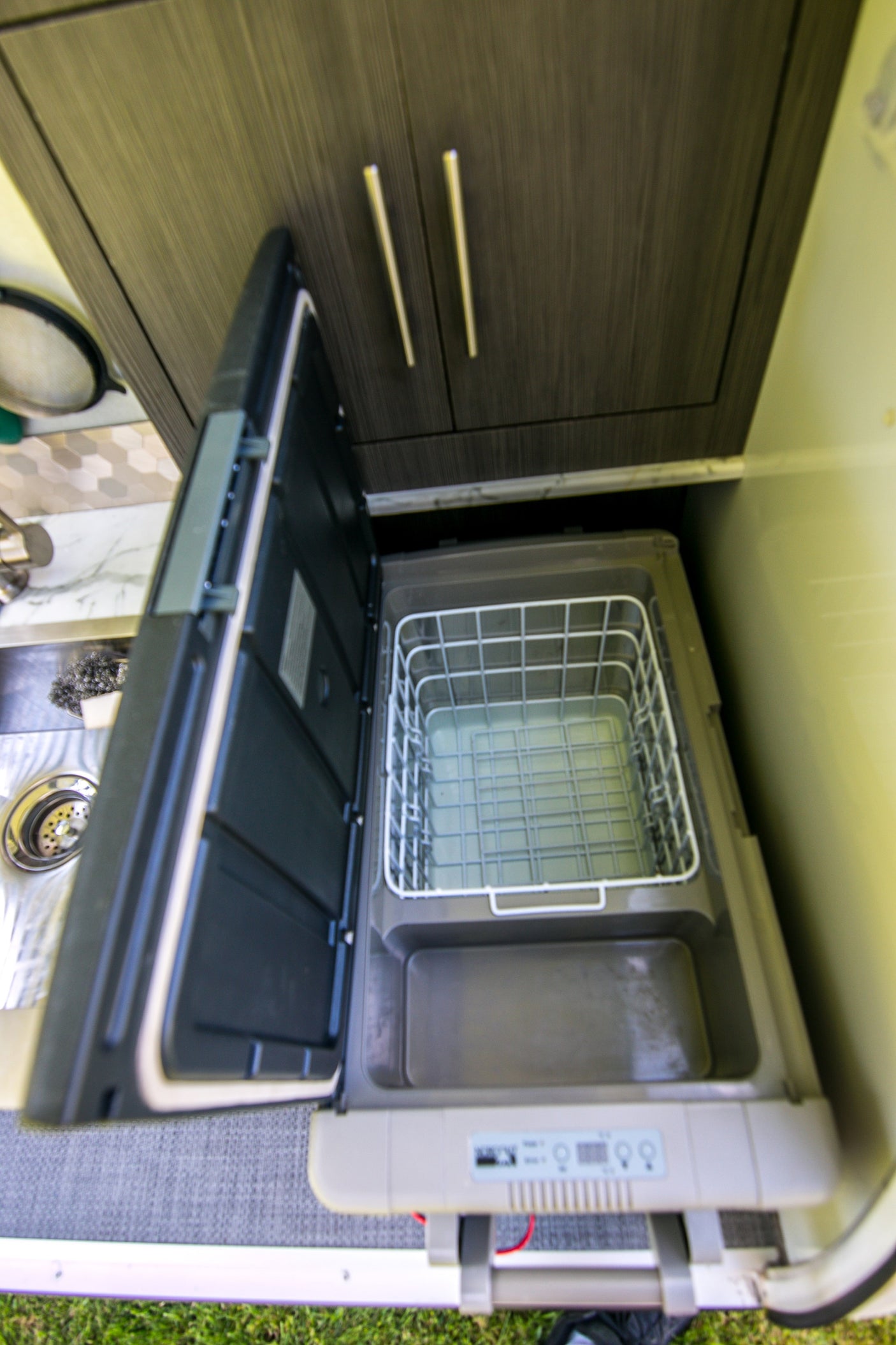 RV rental kitchen: Open portable fridge/freezer with digital controls, alongside a small sink and modern wood-grain cabinets.