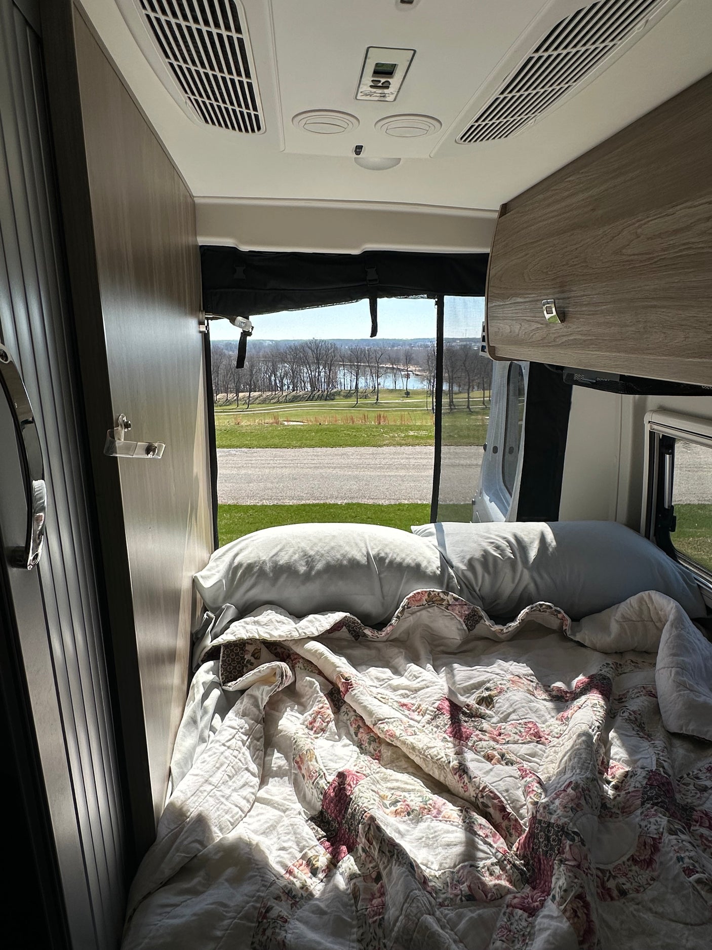 Inside an RV rental: a sunlit bed with floral quilt, offering a scenic view of a lake through bare trees. Perfect for adventure.