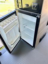 **RV rental** interior features: open, empty mini-fridge with egg holder, control panel (AC/DC/USB outlets, "Coach Batt" switch).