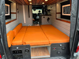 Bright RV rental camper van interior. Features a versatile orange cushioned bed/lounge, wood ceiling, and compact kitchenette.
