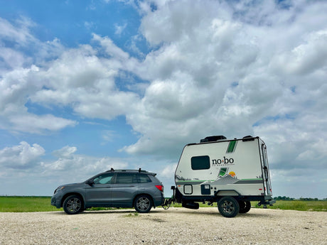 Grey SUV pulling a 'No Bo' RV rental on a gravel road. Ready for outdoor adventure and camping under a bright, cloudy sky.