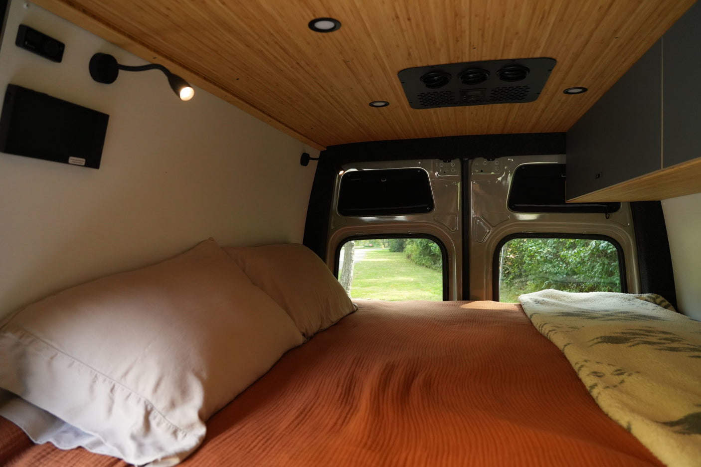 Cozy bed in an RV rental, featuring a wooden ceiling, wall-mounted lights, and scenic views from the back windows.