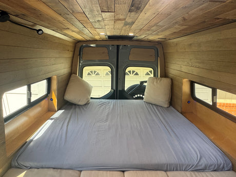 Cozy RV rental camper van interior featuring a comfy bed with light sheets, warm wood paneling on walls & ceiling.
