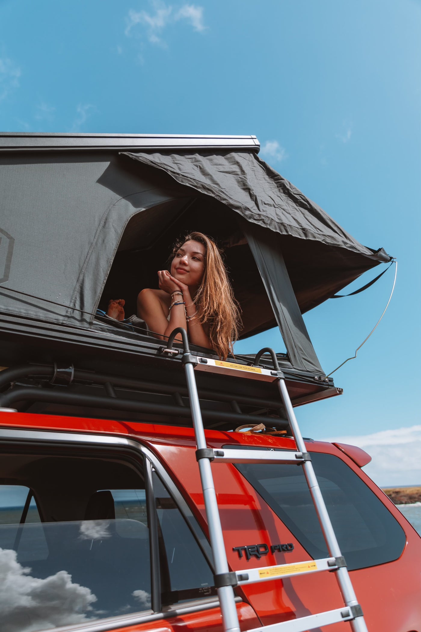 A woman smiles from her rooftop tent on a red TRD PRO RV rental, enjoying the clear sky and scenic outdoor adventure. — photo 5