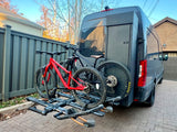 A dark gray rv rental van is parked on pavers, featuring a red and a black mountain bike secured on a rear hitch rack.