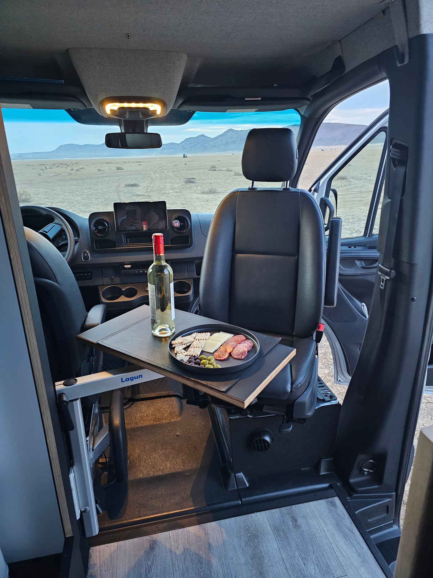 Inside a comfy RV rental: charcuterie plate & wine on a table, with scenic desert mountains visible through the windshield.