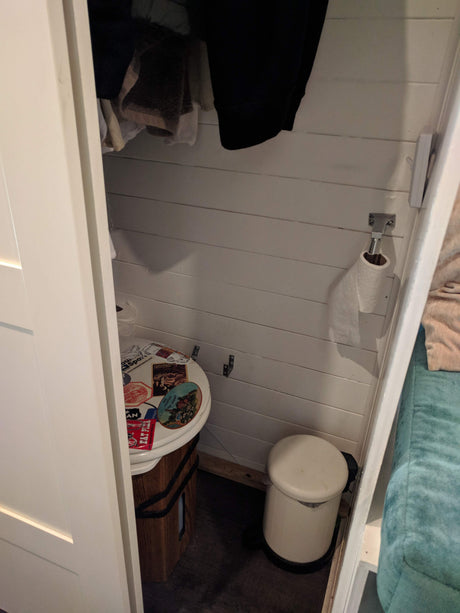 Inside a compact RV rental, a toilet with travel stickers, toilet paper holder, and small trash can are visible.