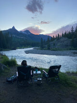 Relaxing riverside with an RV rental view. Two chairs face a scenic mountain and river at sunset, perfect for unwinding.
