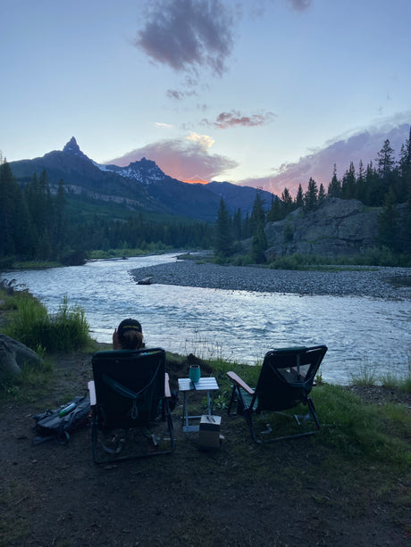 Relaxing riverside with an RV rental view. Two chairs face a scenic mountain and river at sunset, perfect for unwinding.