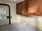 RV rental bedroom setup with a grey bed, wooden cabinets, and a colorful patterned curtain.