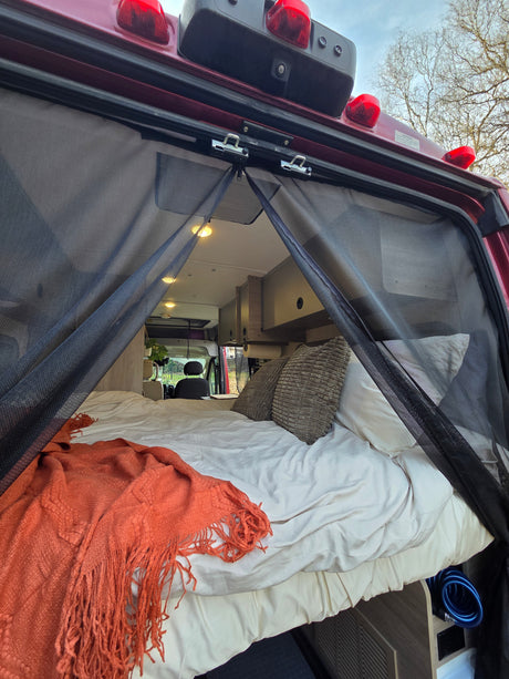 Relax in this RV rental! View of a cozy bed with an orange blanket and bug screen from the open rear of a camper van.
